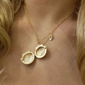 May include: A gold necklace with a seashell pendant that opens. The pendant is open and the inside is visible. The necklace also has a small charm with the letter 'A' on it.