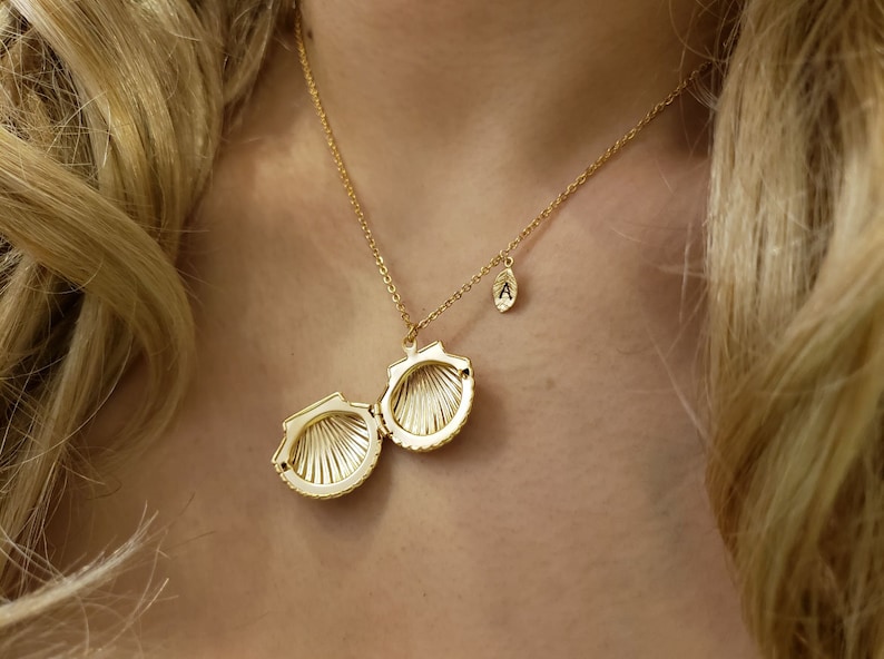 May include: A gold necklace with a seashell pendant that opens. The pendant is open and the inside is visible. The necklace also has a small charm with the letter 'A' on it.