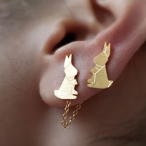 以下が含まれることがあります： つながったチェーンが付いた金色のウサギのイヤリングのペア。イヤリングは人の耳につけられています