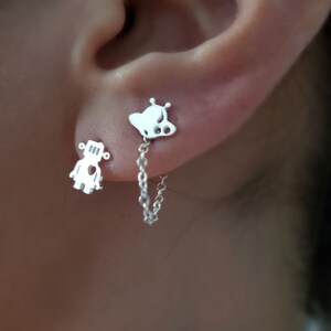 May include: A pair of silver earrings featuring a robot and a butterfly design. The earrings are connected by a silver chain.
