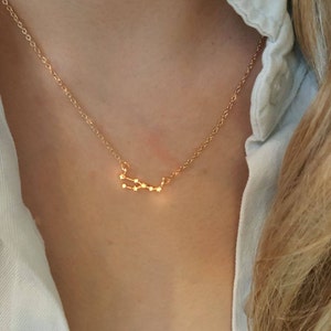 May include: A delicate gold-colored necklace featuring a constellation design. The pendant showcases a series of small, connected dots, forming a celestial pattern. The necklace is worn against a white shirt.