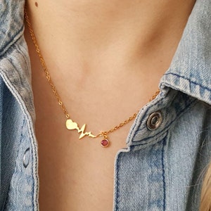 May include: Gold-coloured necklace with a heart, a heartbeat line, and a small red gemstone pendant. The necklace is worn against a denim jacket.