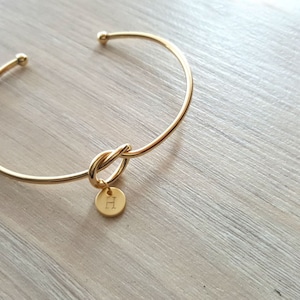 Puede incluir: Un brazalete de oro con un detalle de nudo y un pequeño dije con la letra "H" grabada.