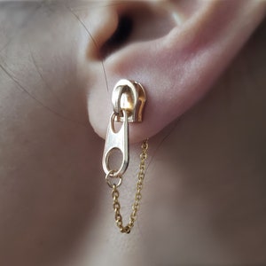 May include: Gold zipper charm earrings with a dangling chain. The zipper charm is silver and the chain is gold.