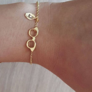 以下が含まれることがあります： 人の手首に2つの金のブレスレット。1つは眼鏡の形をしたチャームが付いたチェーン。もう1つは小さな丸い金のディスクが付いたチェーンです。