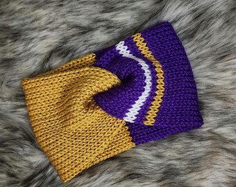 May include: A knitted headband in gold and purple with white and gold accents. The headband is a twisted design, with a solid gold section on one side and a purple section with a white and gold wave pattern on the other.