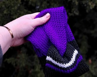 May include: A knitted headband in shades of purple, black, and white. The headband features a chevron design in white and black against a black base, transitioning to a solid purple. The item is held up against a blurred green background.