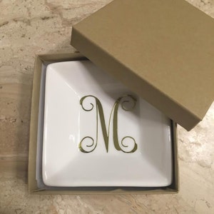 May include: A white ceramic trinket dish with a gold 'M' monogram. The dish is in a brown cardboard box.