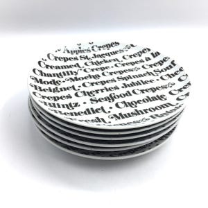 May include: A stack of six white ceramic dessert plates with black text listing different types of crepes. The text includes "Apples Crepes", "Crepes St. Jacques", "Crepes à la Mode", "Mocha Crepes", "Crepes Spinach Souf", "Beignet", "Cherries Jubilee", "Seafood Crepes", "Blintz", "Benedict", "Chocolate Crepes", "Mushroom Crepes", and "Fresh Crepes".