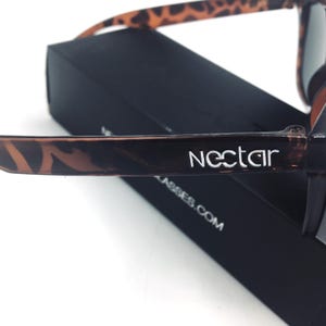 Op de afbeelding: Close-up van een paar zwarte en bruine zonnebrillen met luipaardprint met het woord "nectar" in wit gedrukt aan de zijkant.