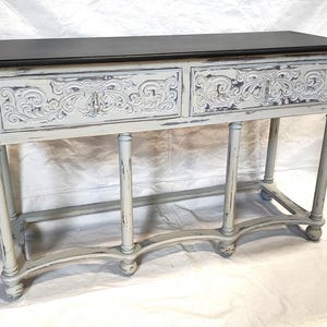 May include: A distressed, off-white console table with a dark gray top. The two drawers feature ornate, carved floral designs. The table has four slender, turned legs and a curved base, showcasing a vintage aesthetic.