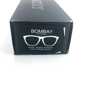 Op de afbeelding: Een zwarte doos met de woorden "Bombay Polarized" en een afbeelding van een zonnebril met bruine schildpad-monturen en oranje crush glazen. De tekst onder de zonnebril luidt "Frame/Brown Tortoise Lens/Orange Crush."
