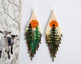 May include: A pair of beaded earrings with a sunset and forest scene. The earrings are made with white, orange, green, and brown beads. The earrings are teardrop shaped and have a fringe.