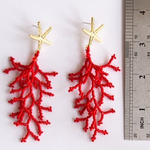 Puede incluir: Un par de pendientes de cuentas de coral rojo con detalles de estrella de mar dorada. Los pendientes miden aproximadamente 5 cm de largo.