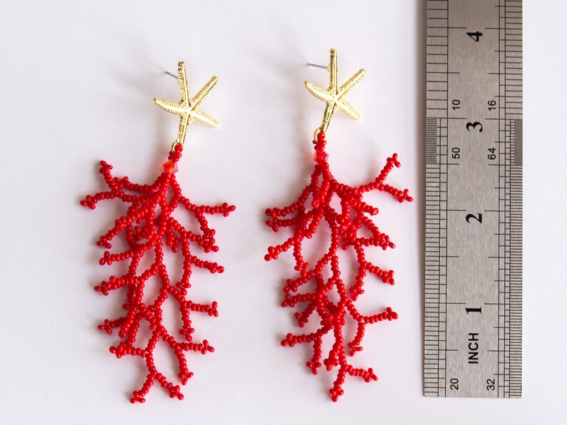 Puede incluir: Un par de pendientes de cuentas de coral rojo con detalles de estrella de mar dorada. Los pendientes miden aproximadamente 5 cm de largo.