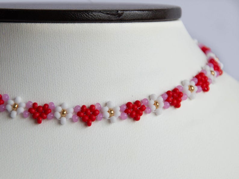 Peut inclure: Un collier ras de cou en perles rouges, blanches et roses avec un motif en forme de c&oelig;ur. Le collier est fait de petites perles et a un aspect d&eacute;licat.