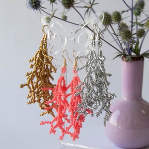 May include: Three pairs of beaded dangle earrings displayed on a butterfly-shaped stand. The earrings are made of gold, silver, and coral-coloured beads.