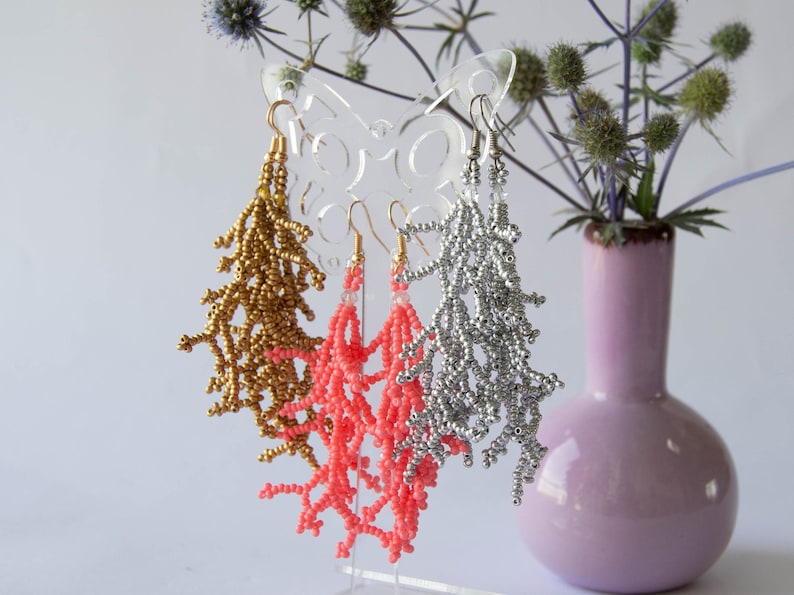 May include: Three pairs of beaded dangle earrings displayed on a butterfly-shaped stand. The earrings are made of gold, silver, and coral-coloured beads.