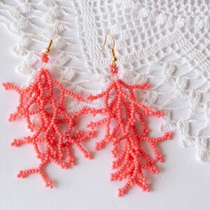 May include: A pair of coral-coloured beaded earrings shaped like coral branches. The earrings are hanging from gold hooks.