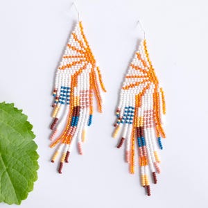 May include: A pair of white beaded earrings with a geometric design featuring orange, yellow, blue, and brown accents. The earrings are long and dangle, with a fringe-like effect.