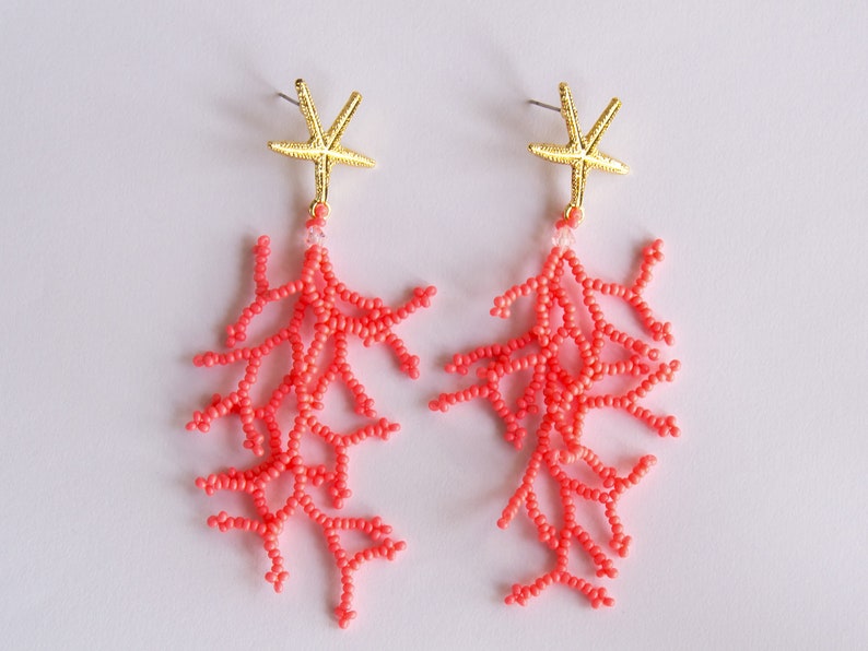Puede incluir: Un par de pendientes de cuentas de color coral con abalorios de estrella de mar dorados. Los pendientes son largos y colgantes, con las cuentas de coral formando un delicado dise&ntilde;o de ramas.