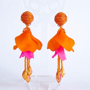 May include: A pair of striking earrings. Each earring has a beaded orange sphere, a large orange petal, a smaller pink petal, and dangling amber beads. These earrings are designed to be a bold and fashionable accessory.