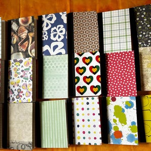 May include: A collection of 16 small notebooks with various patterned covers. The covers feature designs such as seashells, butterflies, hearts, polka dots, and abstract patterns. The notebooks are bound with black spines and have a brown paper cover.