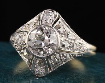 Könnte beinhalten: Ein Vintage-Verlobungsring mit Diamanten, der einen großen runden Mitteldiamanten hat, der von kleineren Diamanten umgeben ist. Der Ring ist in Platin gefasst und hat ein zartes, filigranes Design.