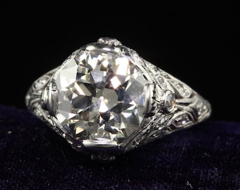 Könnte beinhalten: Ein Vintage-Verlobungsring aus Platin mit einem großen, runden Brillanten, der in einem filigranen Design gefasst ist. Der Ring ist mit kleineren Diamanten verziert.