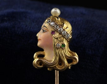 May include: A gold-toned stick pin featuring a profile of a woman with a pearl crown and a face painted in enamel. The woman's hair is detailed with gold and the pin is set with diamonds, rubies, and emeralds.