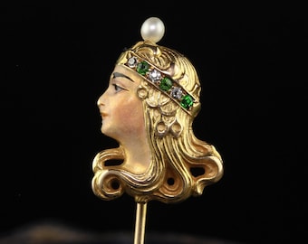 May include: A gold-toned brooch featuring a woman's profile with a pearl crown and green and white gemstones. The brooch is attached to a gold pin.