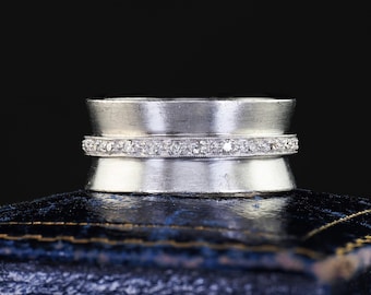 Puede incluir: Un anillo de banda ancha de plata con una fila horizontal central de pequeñas piedras claras de corte redondo engastadas en un delicado borde milgrain. La superficie metálica tiene un acabado mate cepillado que contrasta con las piedras.