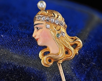May include: A vintage gold-tone hat pin featuring a profile of a woman's face. The woman has long, flowing golden hair, a pearl on top, and a jeweled headband. The face is painted with delicate features and a rosy complexion. A small green gem is on the hair.