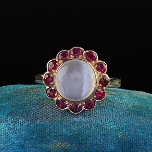 Op de afbeelding: Een gouden ring met een grote, ronde, lichtblauwe edelsteen in het midden, omringd door kleinere, rode edelstenen. De ring heeft een eenvoudige, gepolijste gouden band. De ring ligt op een turquoise achtergrond.