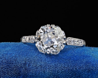 Puede incluir: Un anillo de plata con un gran diamante de talla cojín transparente engastado en un engaste delicado y ornamentado. La banda está acentuada con diamantes más pequeños.