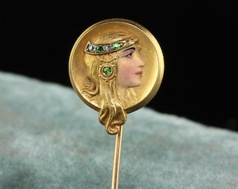 May include: A gold-toned stick pin featuring a profile of a woman with long, flowing hair. The woman's face is painted with delicate features. The pin is adorned with green and white gemstones, adding a touch of sparkle to this antique jewelry piece.