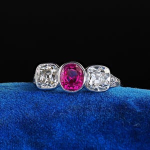 May include: A platinum ring featuring three gemstones: a central oval-cut ruby flanked by two square-cut diamonds. The ruby is a vibrant pink, and the diamonds are clear. The ring is set on a blue surface against a black background.