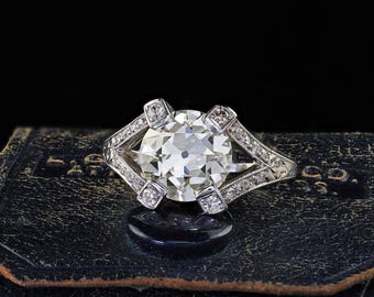 Könnte beinhalten: Ein Platinring mit einem großen, runden Diamanten im Brillantschliff als Mittelpunkt. Der Ring hat kleinere Diamanten, die entlang des Bandes und in dekorativen Fassungen um den Hauptstein gesetzt sind. Der Ring wird auf einer dunkelblauen Lederoberfläche präsentiert.