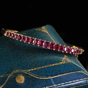 May include: A hinged bracelet featuring a continuous row of rectangular, deep red rubies set in a gold metal frame. Small, clear, round accent stones are positioned between each ruby, creating a sparkling, linear design across the entire band.