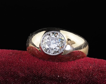 May include: A gold ring with a large, round, clear diamond set in the center. The ring is on a red velvet surface, with a black background. The ring's band is thick and polished.