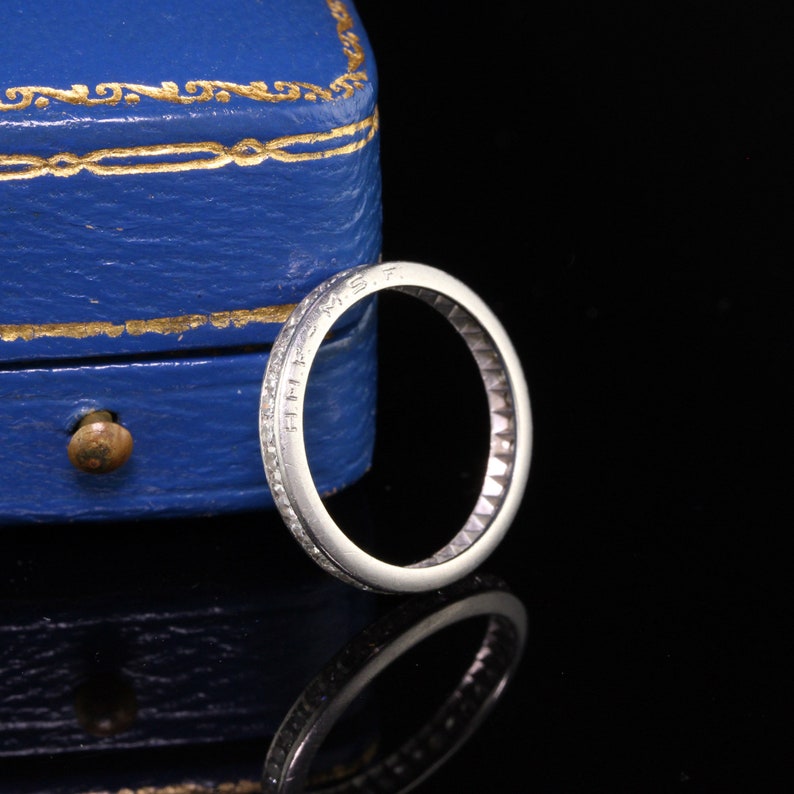 May include: A thin silver band with small diamonds set in a channel setting. The band is inscribed with the letters "E.M.S.T."