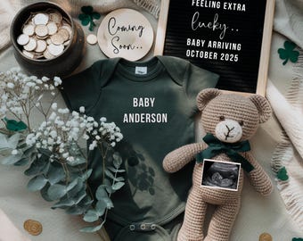 Op de afbeelding: Een groene baby romper met de tekst "Baby Anderson" erop. Een gehaakte bruine teddybeer met een groen lint en een echobeeld op zijn borst. Een houten bord met de tekst "Feeling extra lucky... Baby arriving October 2025".