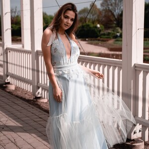 May include: A light blue floor-length halter dress featuring a deep V-neckline, intricate white floral embroidery on the bodice, and a layered tulle skirt overlay that creates a soft, flowing silhouette with sheer fabric panels.