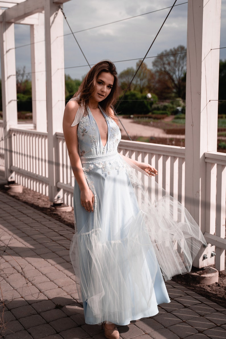 May include: A light blue floor-length halter dress featuring a deep V-neckline, intricate white floral embroidery on the bodice, and a layered tulle skirt overlay that creates a soft, flowing silhouette with sheer fabric panels.