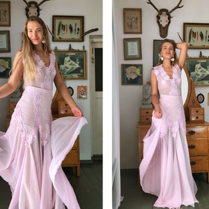 May include: A woman wearing a light pink, floor-length dress with lace detailing and a sheer overlay. The dress has a V-neckline and a fitted bodice. The woman is standing in a room with a vintage aesthetic, and she is holding the skirt of the dress in her hands.