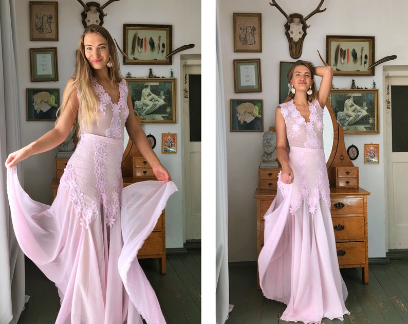 May include: A woman wearing a light pink, floor-length dress with lace detailing and a sheer overlay. The dress has a V-neckline and a fitted bodice. The woman is standing in a room with a vintage aesthetic, and she is holding the skirt of the dress in her hands.