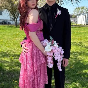K&ouml;nnte beinhalten: Ein Paar posiert f&uuml;r ein Foto, die Frau in einem rosafarbenen, mehrlagigen Spitzenkleid und der Mann im schwarzen Anzug. Die Frau h&auml;lt einen Strau&szlig; rosa und wei&szlig;er Blumen.