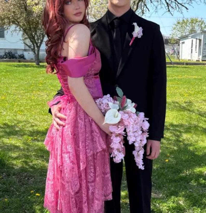 K&ouml;nnte beinhalten: Ein Paar posiert f&uuml;r ein Foto, die Frau in einem rosafarbenen, mehrlagigen Spitzenkleid und der Mann im schwarzen Anzug. Die Frau h&auml;lt einen Strau&szlig; rosa und wei&szlig;er Blumen.