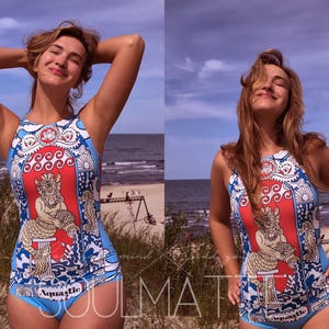 Può includere: Un costume da bagno intero blu e bianco con una grafica di sirena e la scritta "Aquastic". Il costume da bagno ha una scollatura alta e un fondo sgambato. Lo sfondo è una scena di spiaggia con il mare e il cielo.