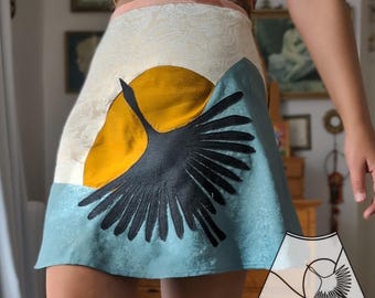 May include: A blue, yellow, and white mini skirt with a black crane design. The skirt is made of a silky fabric and has a high waist.